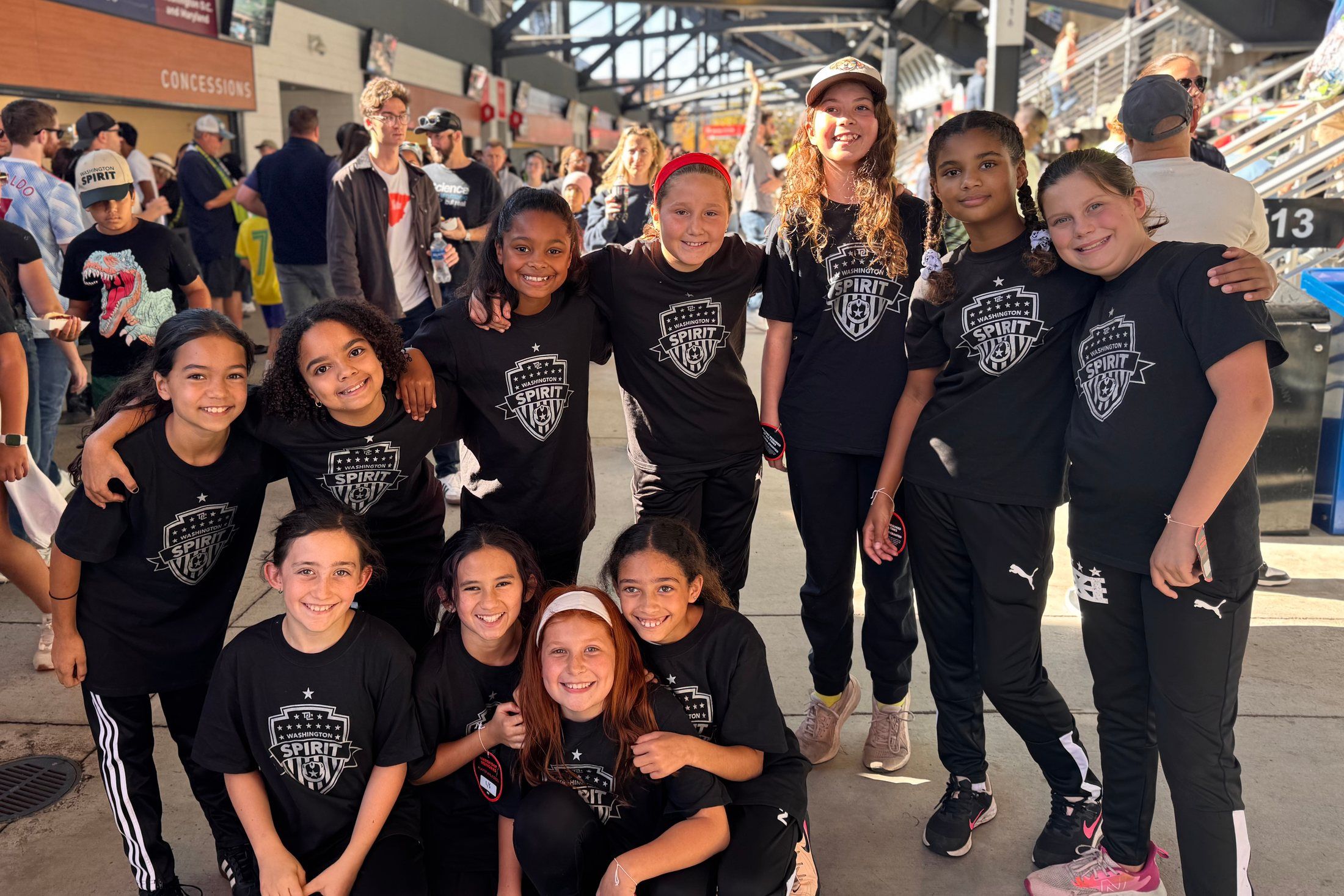 A group of ten smiling girls in matching black team shirts, standing and crouching with their arms around each other.