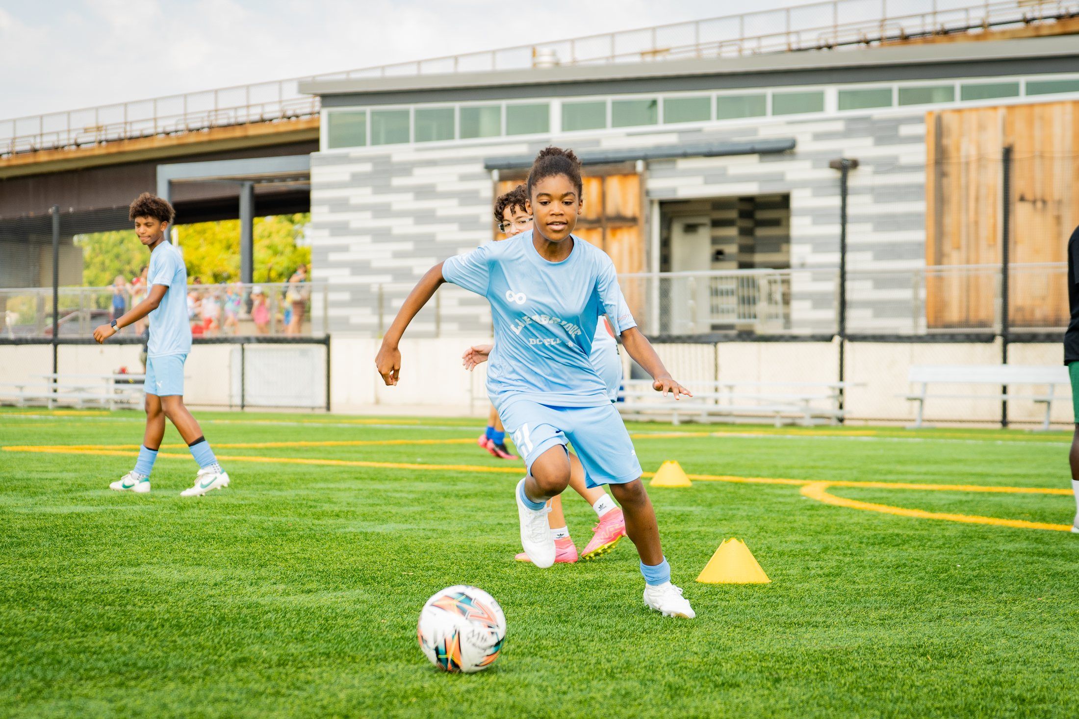 A girl in a light blue shirt dribbles a soccer ball past yellow cones on a sunny field.