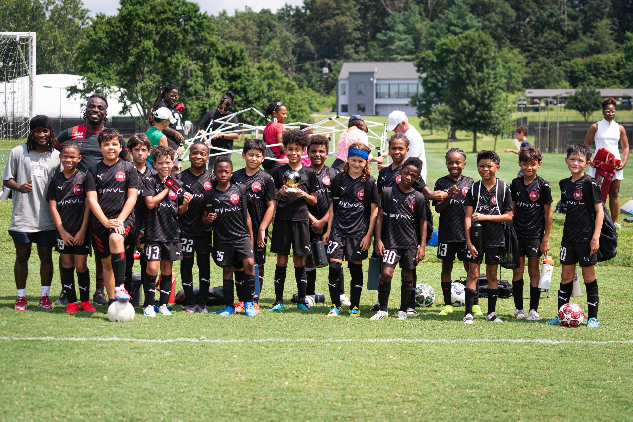 A youth soccer team in black kits lined up on a grass field with their coaches, one player holding a small trophy.