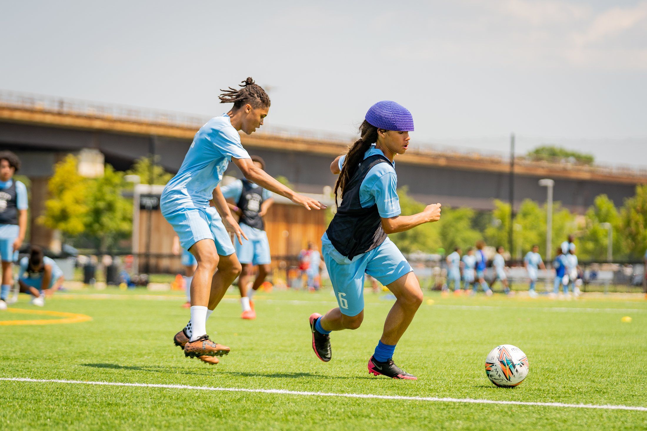 Two teenage players in light blue kits chase a soccer ball on a sunny field.