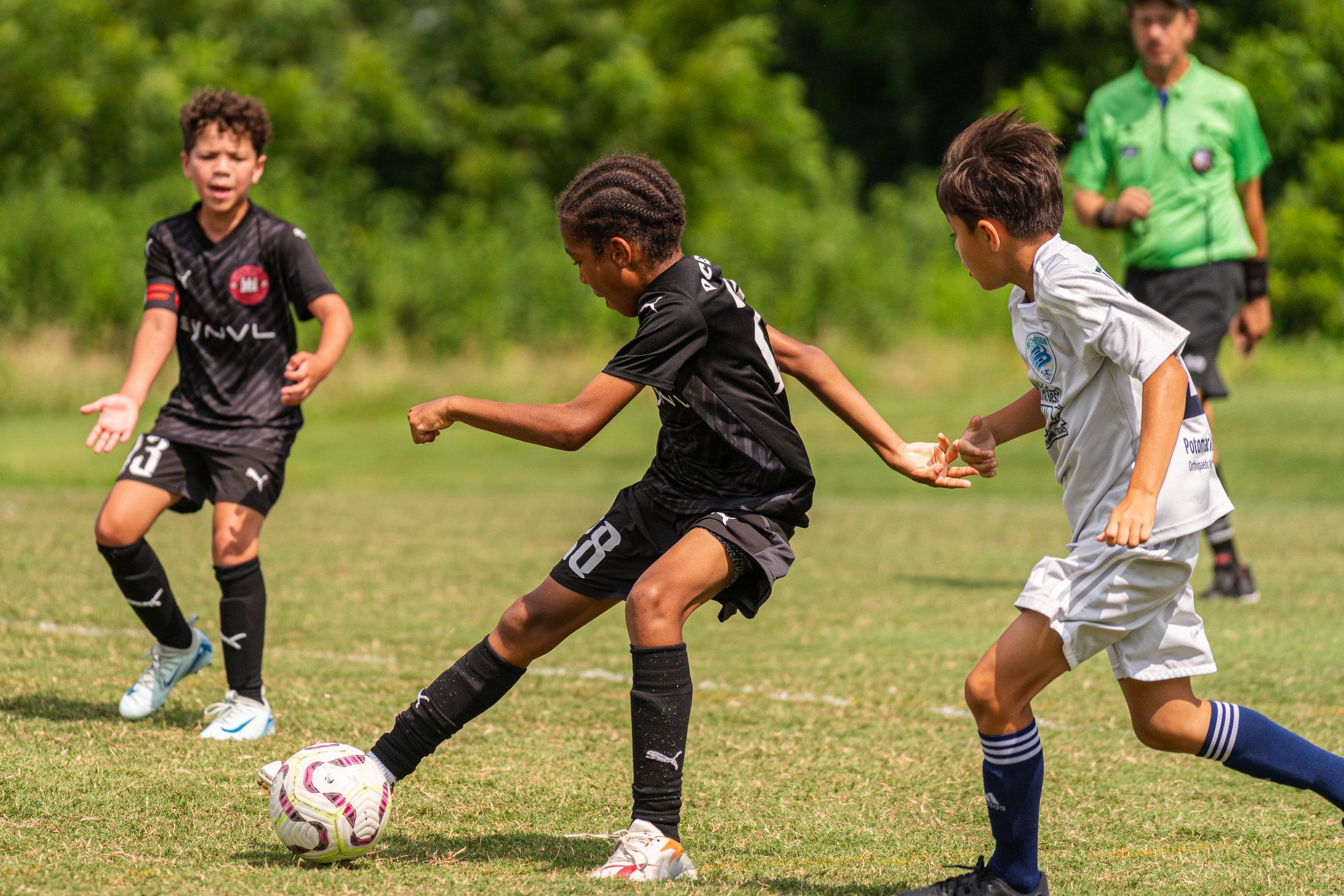 A player in a black kit kicks a soccer ball as a teammate and an opponent in a white kit run alongside on a grass field.