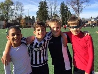 2015: Four boys with their arms around each other, smiling, on an artificial turf field.