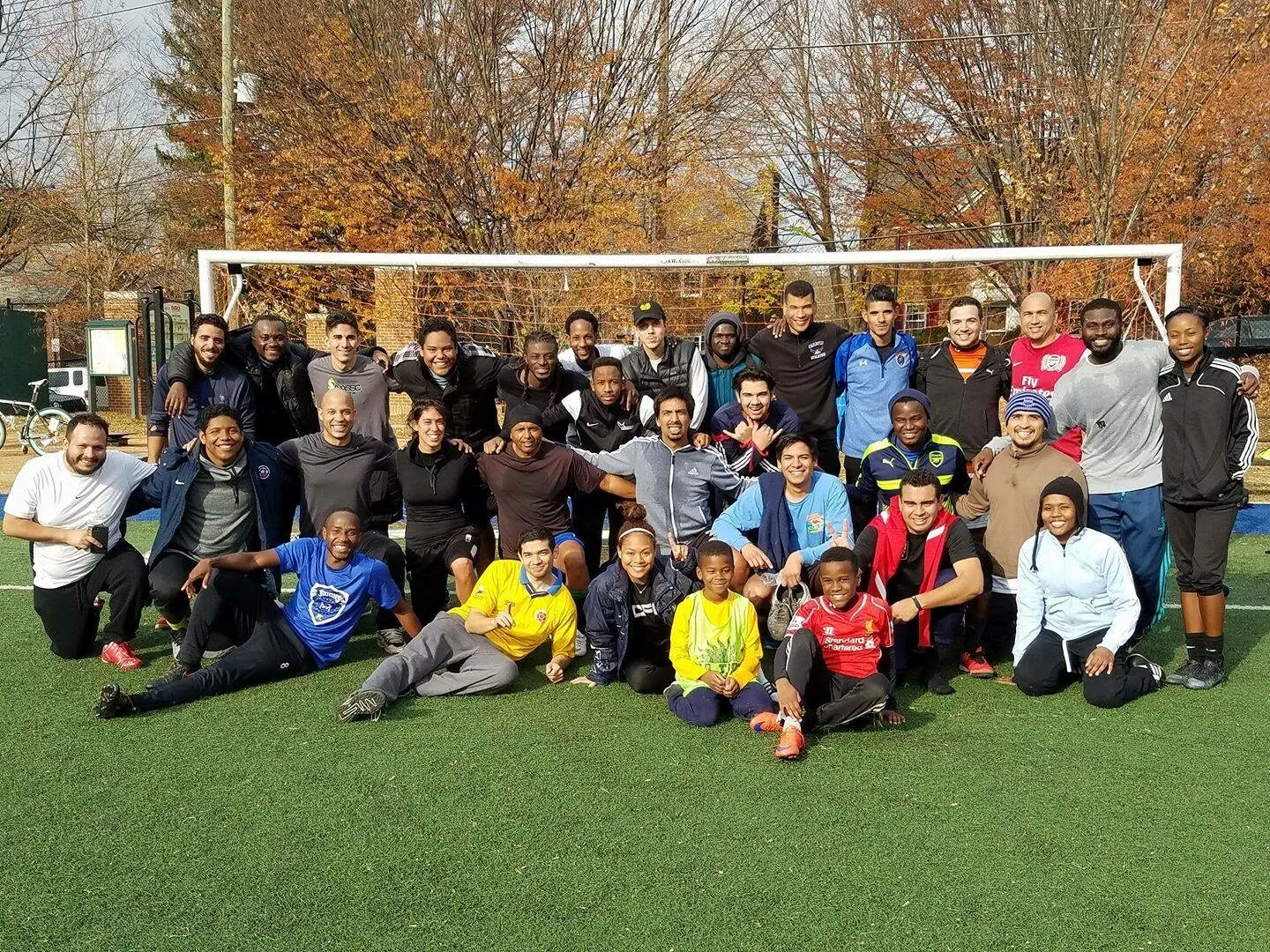 2016: A large group of adults and children pose together in front of a goal on a sunny autumn day.