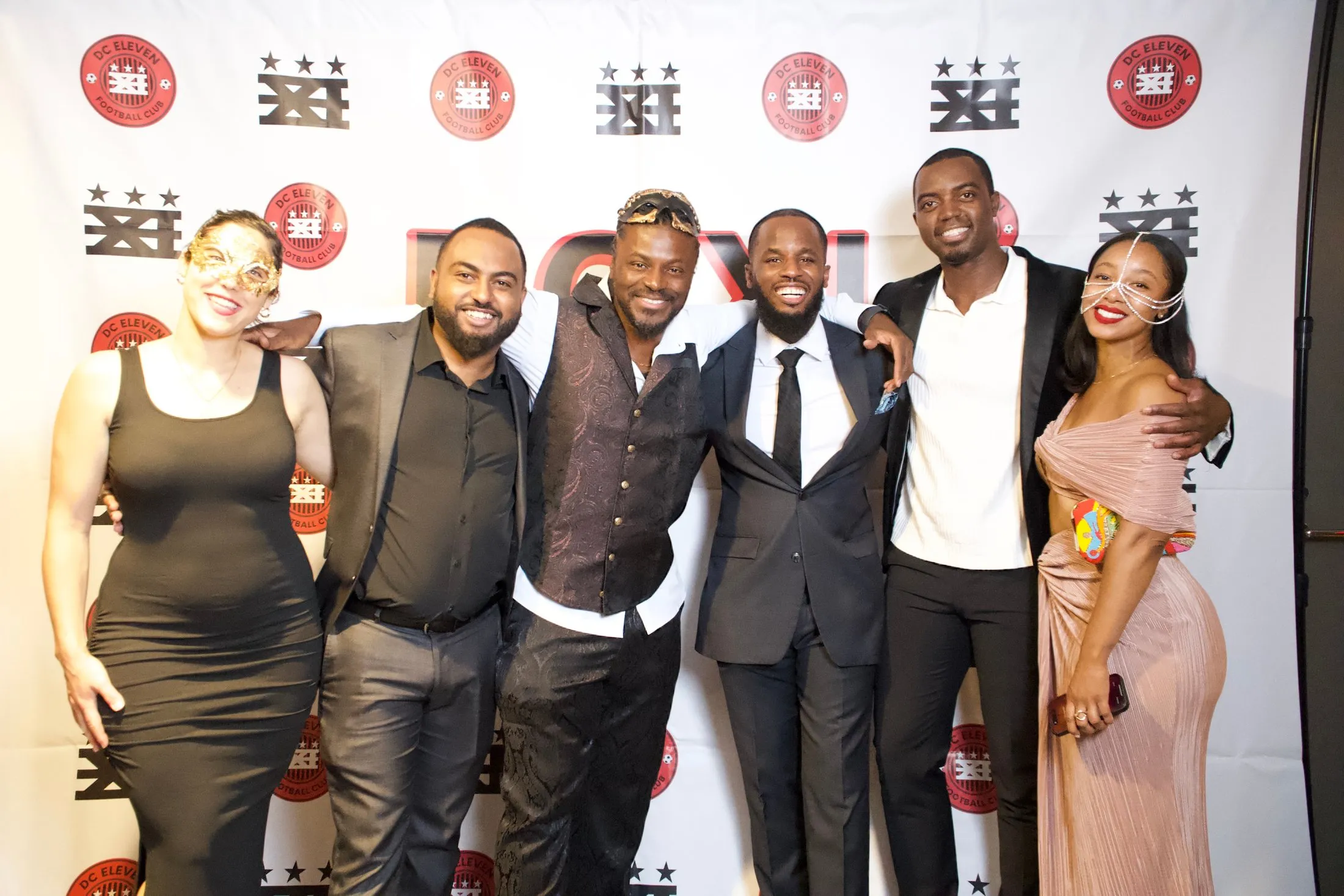 Six gala guests in evening wear pose arm in arm in front of a DC XI backdrop.