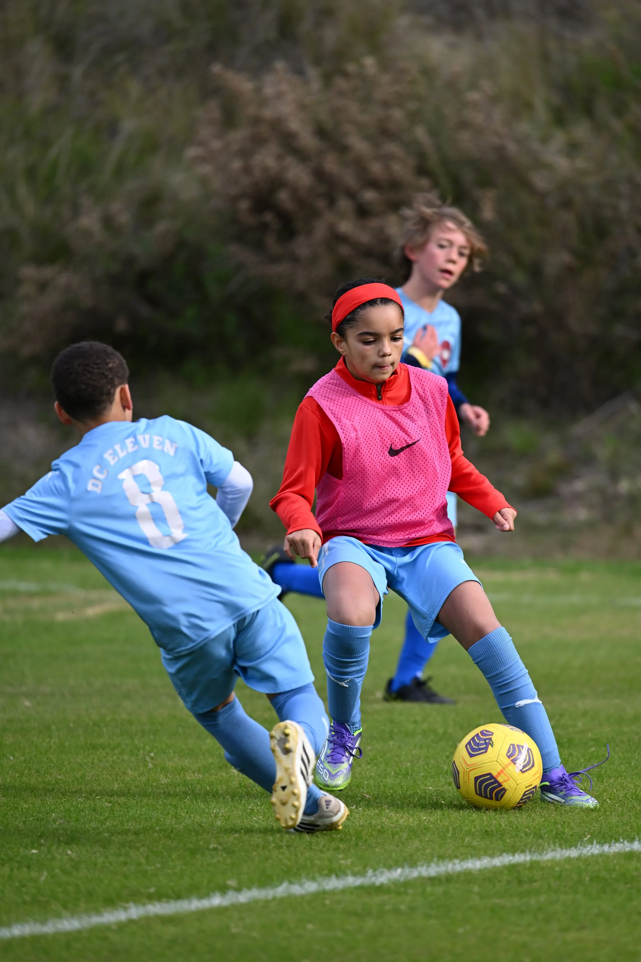 Two young players, one in a pink bib and red headband, challenge for a yellow ball.