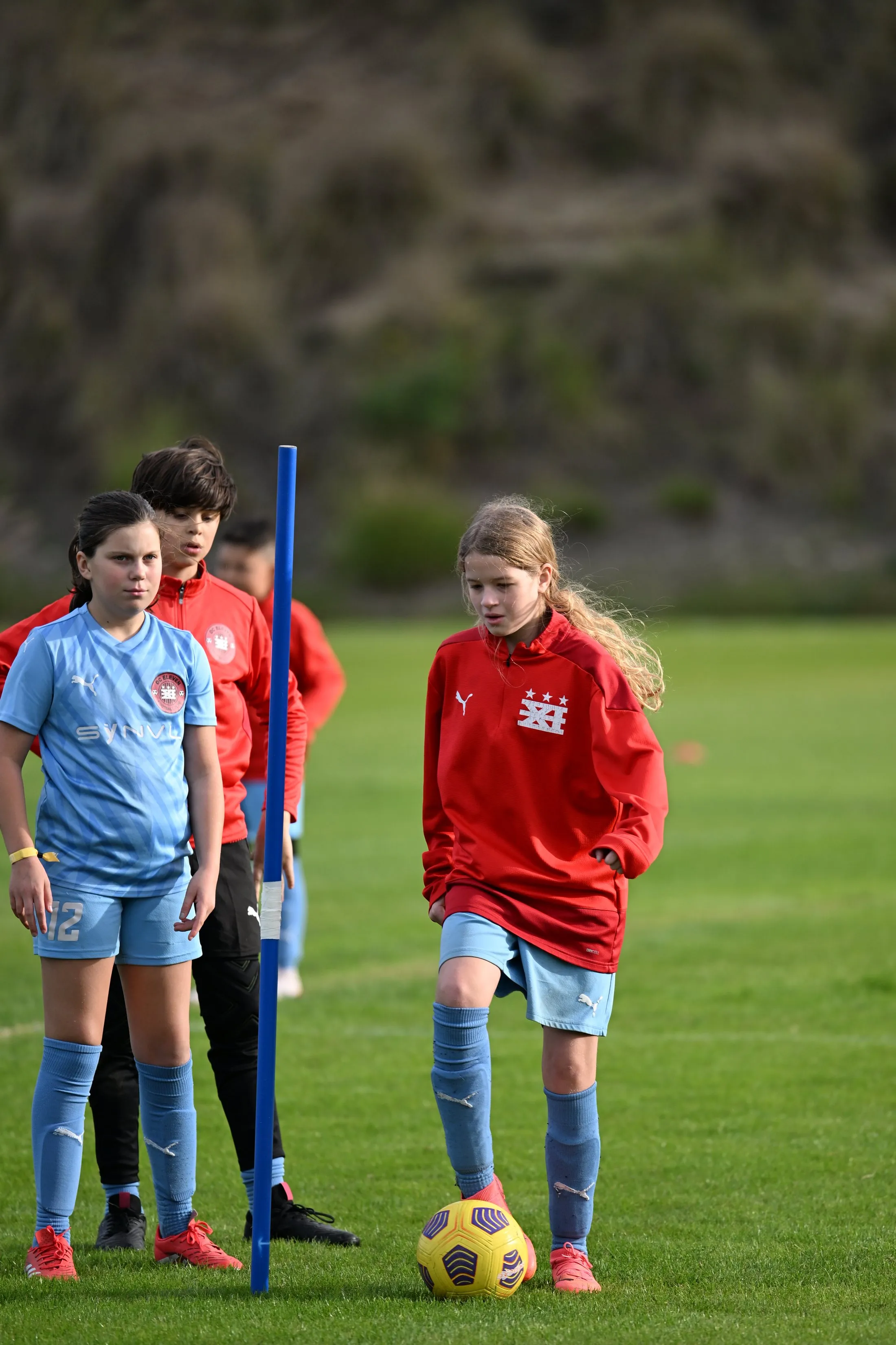 Two girls in light blue and red training tops take part in a dribbling drill.