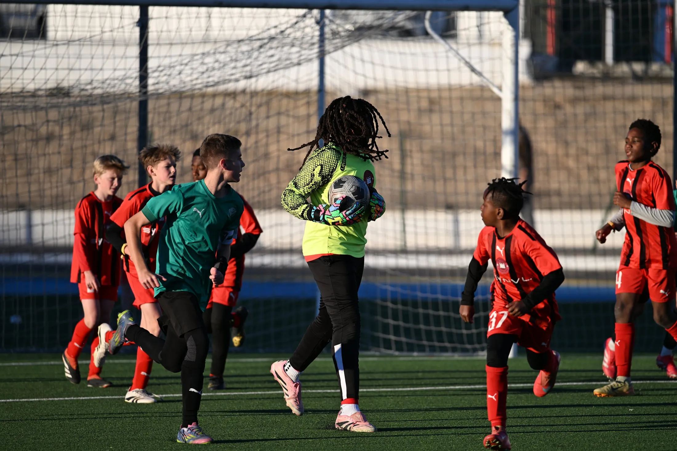 A goalkeeper catches the ball while players in red rush the goal.