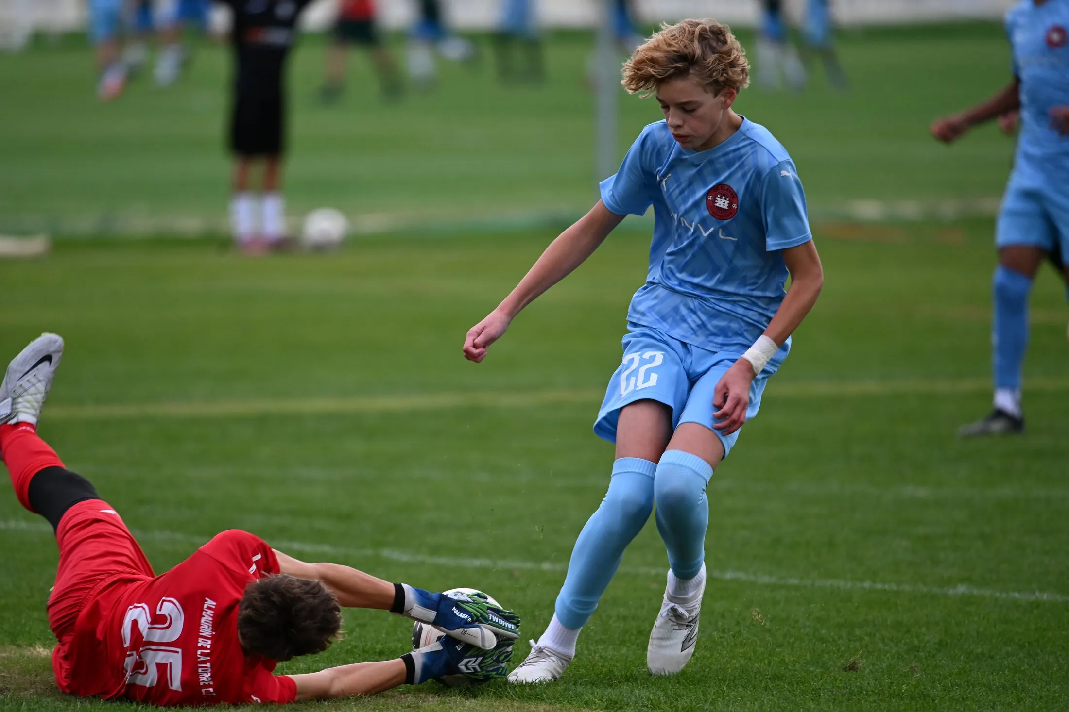 A goalkeeper dives to block the ball at the feet of an attacker in a light blue kit.