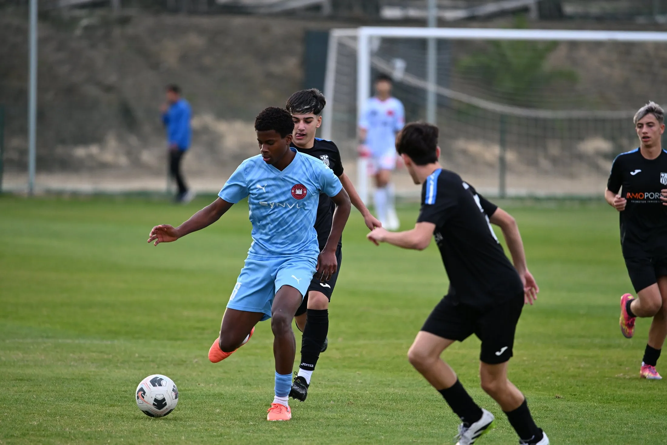 An older player in a light blue kit dribbles past a defender in black during a match.