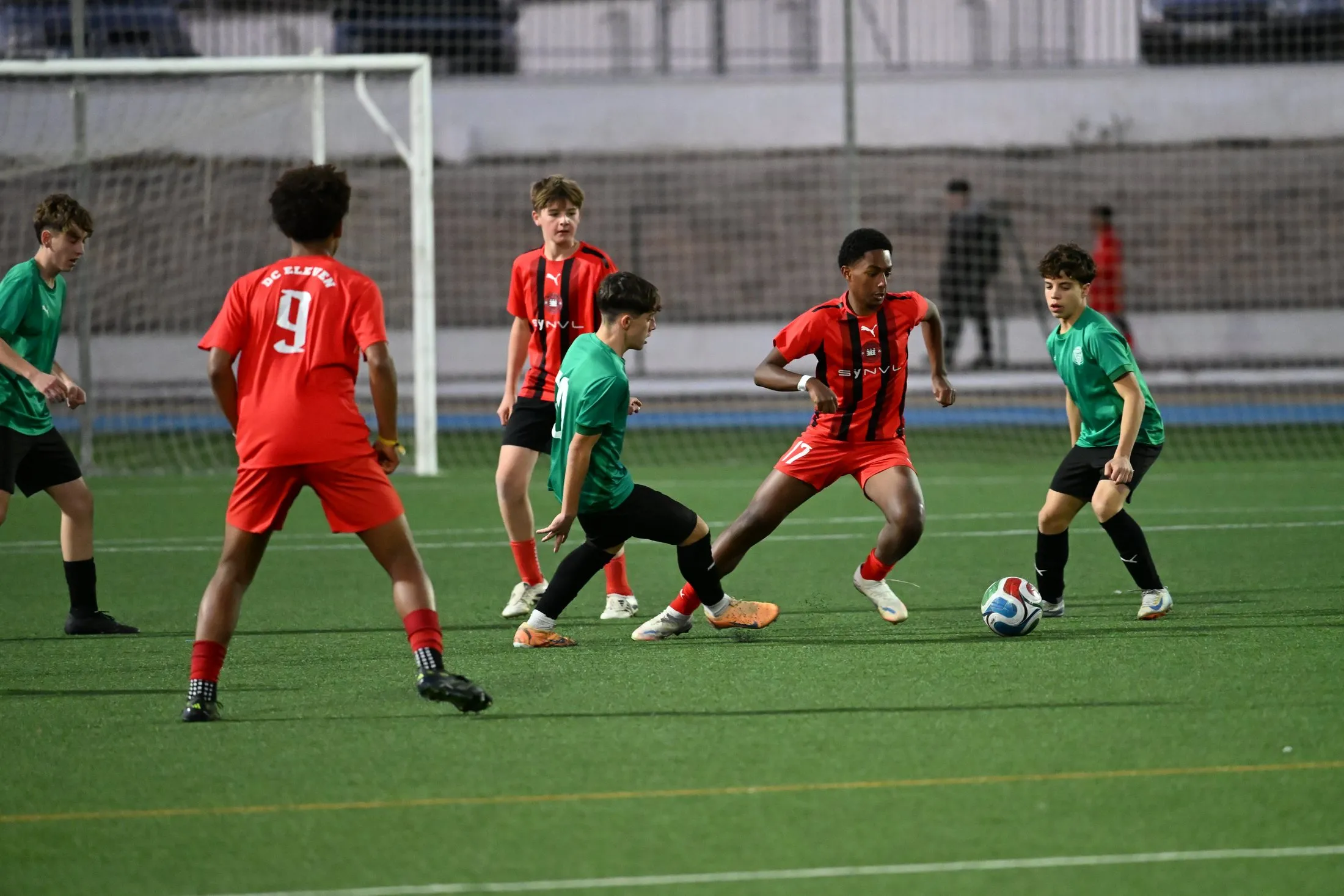 Players in red and black and players in green contest the ball in a match on an artificial pitch in Spain.