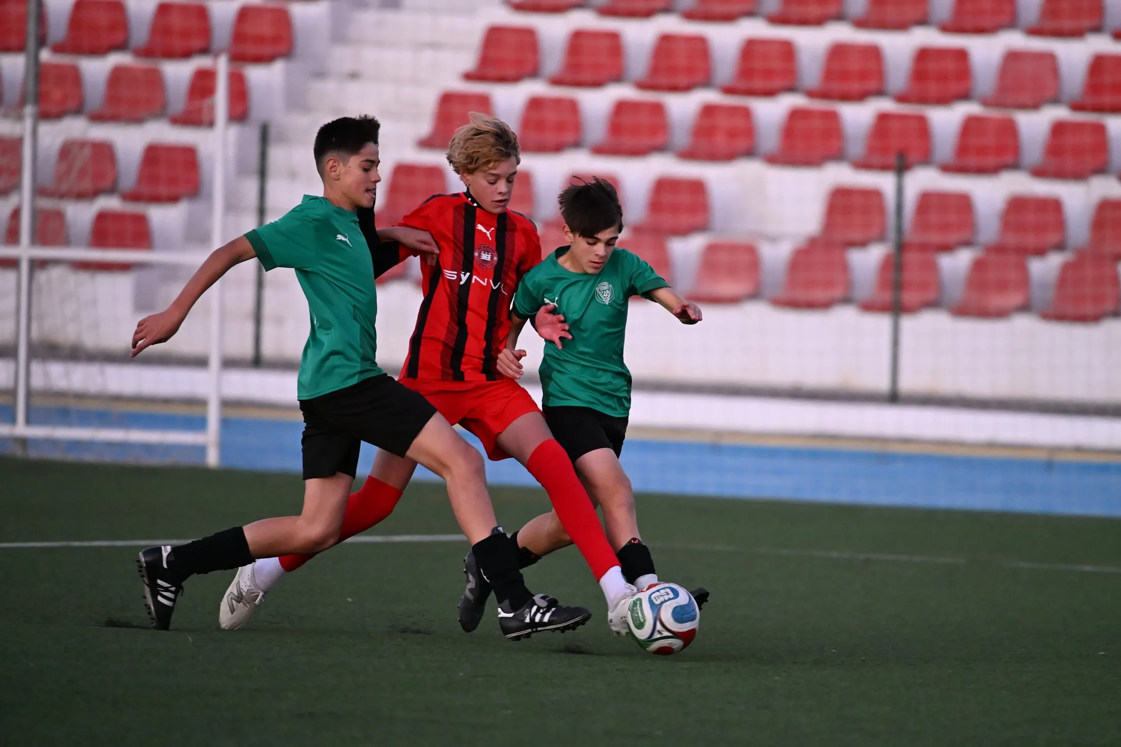A player in red is challenged by two opponents in green in front of empty red stadium seats.