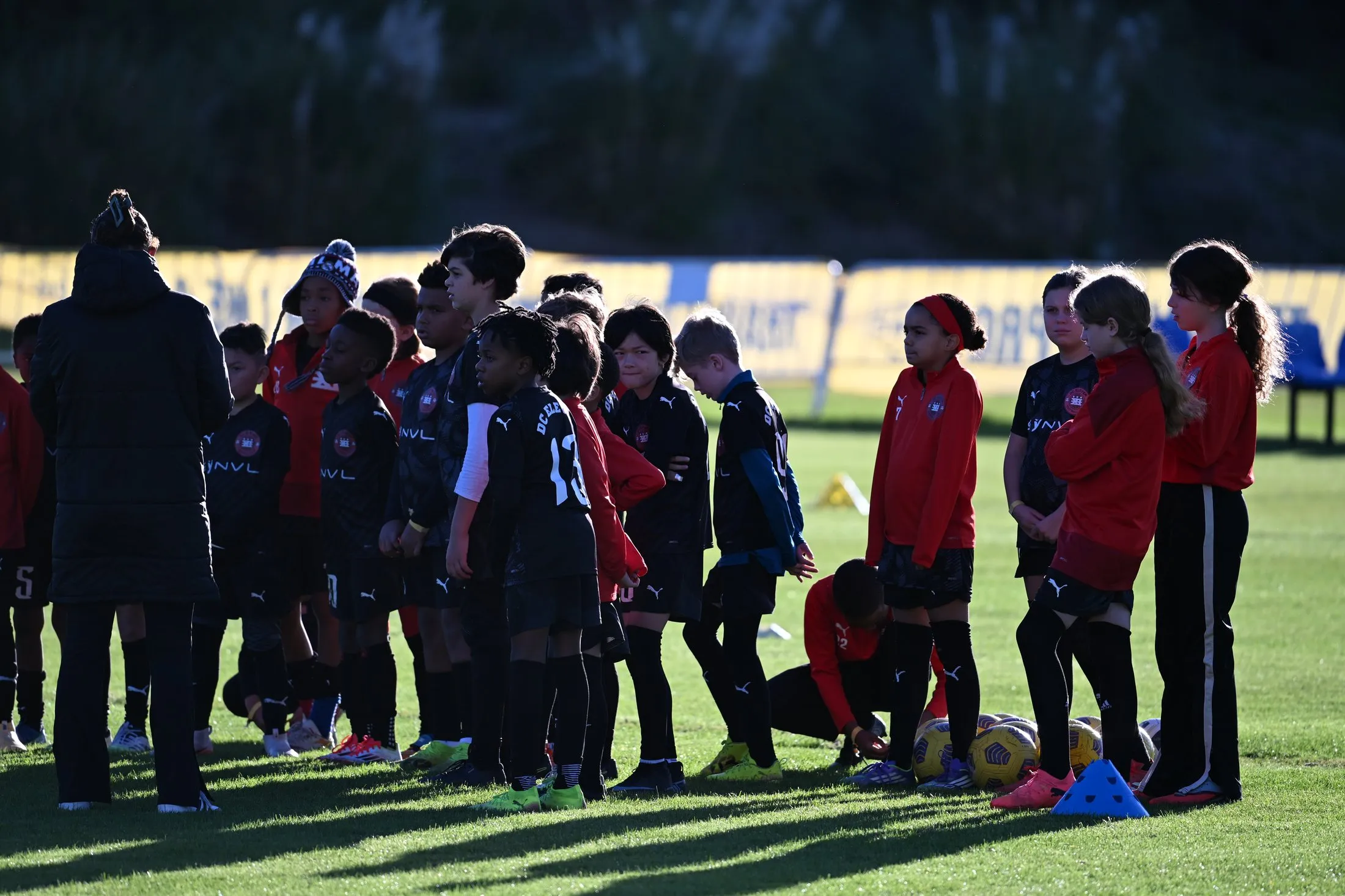 A mixed group of young players in DC Eleven kit stand in a line listening to coaches on a pitch.
