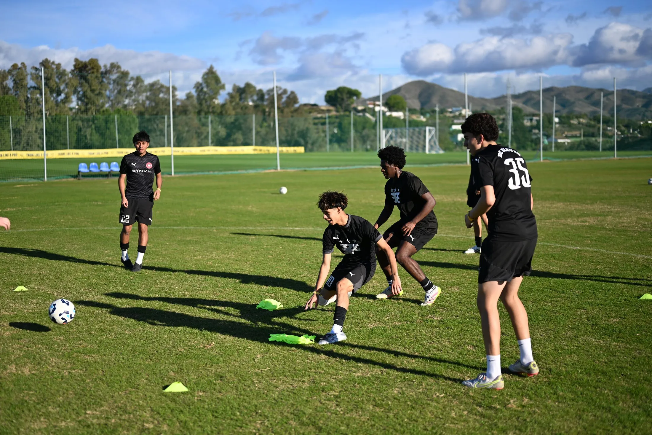 Players in black training kit work on a ball drill on a grass pitch with mountains in the background.