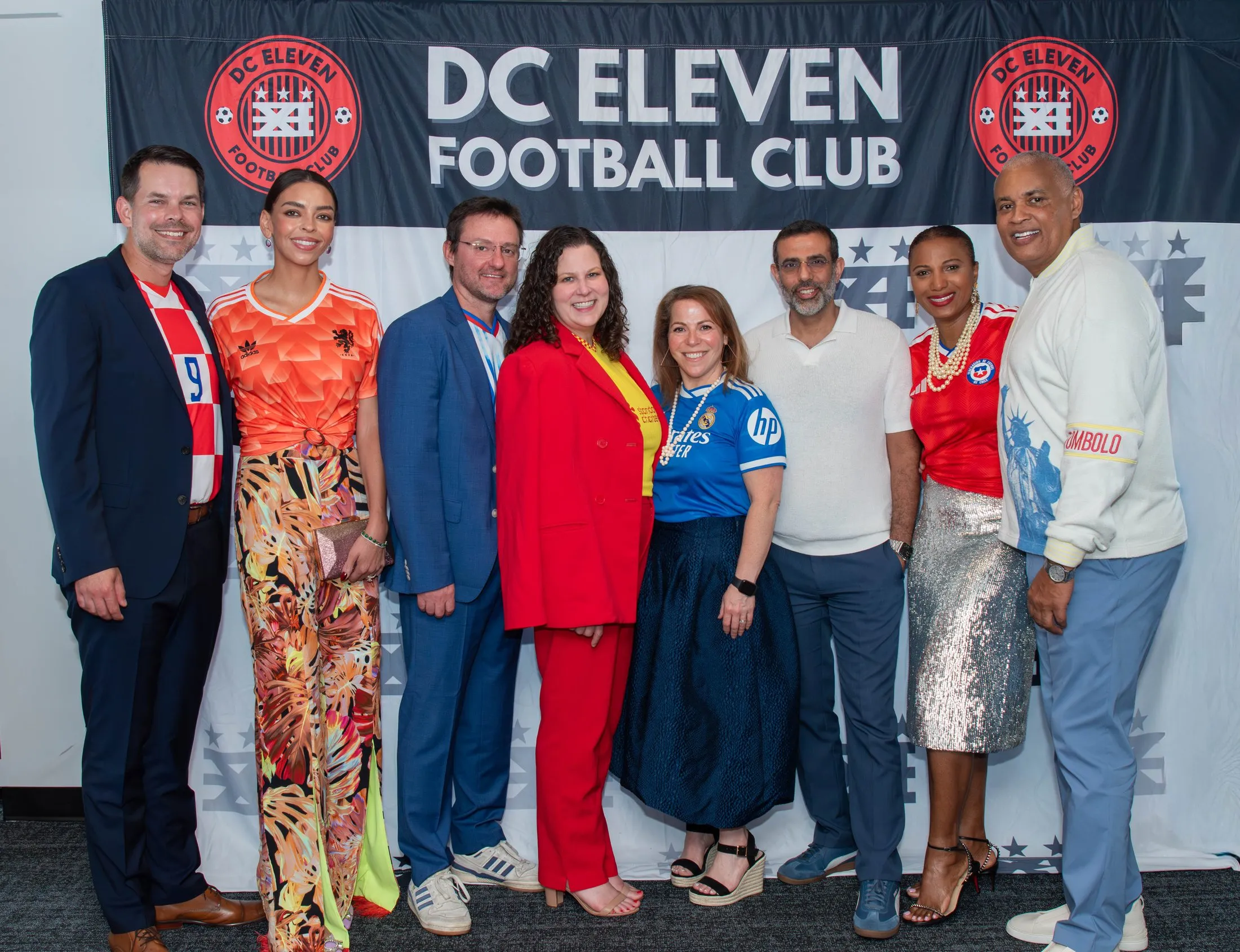 Seven gala guests, women and men, standing together in front of a DC Eleven Football Club banner, several wearing football shirts.