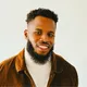 AJ Nwosu, smiling, with a beard, wearing a brown jacket over a white turtleneck