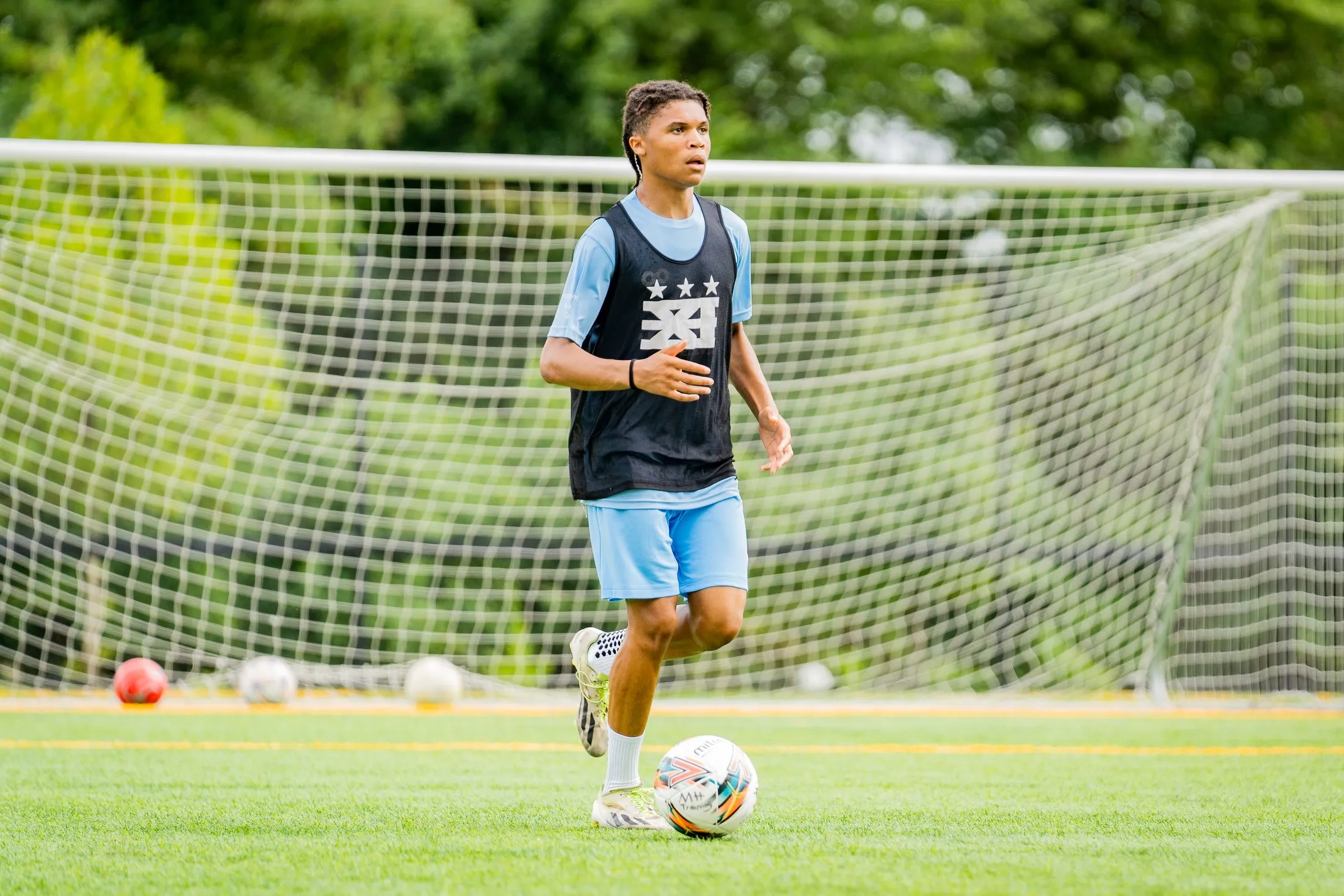 A player in a black bib dribbles a ball in front of a goal net during training.