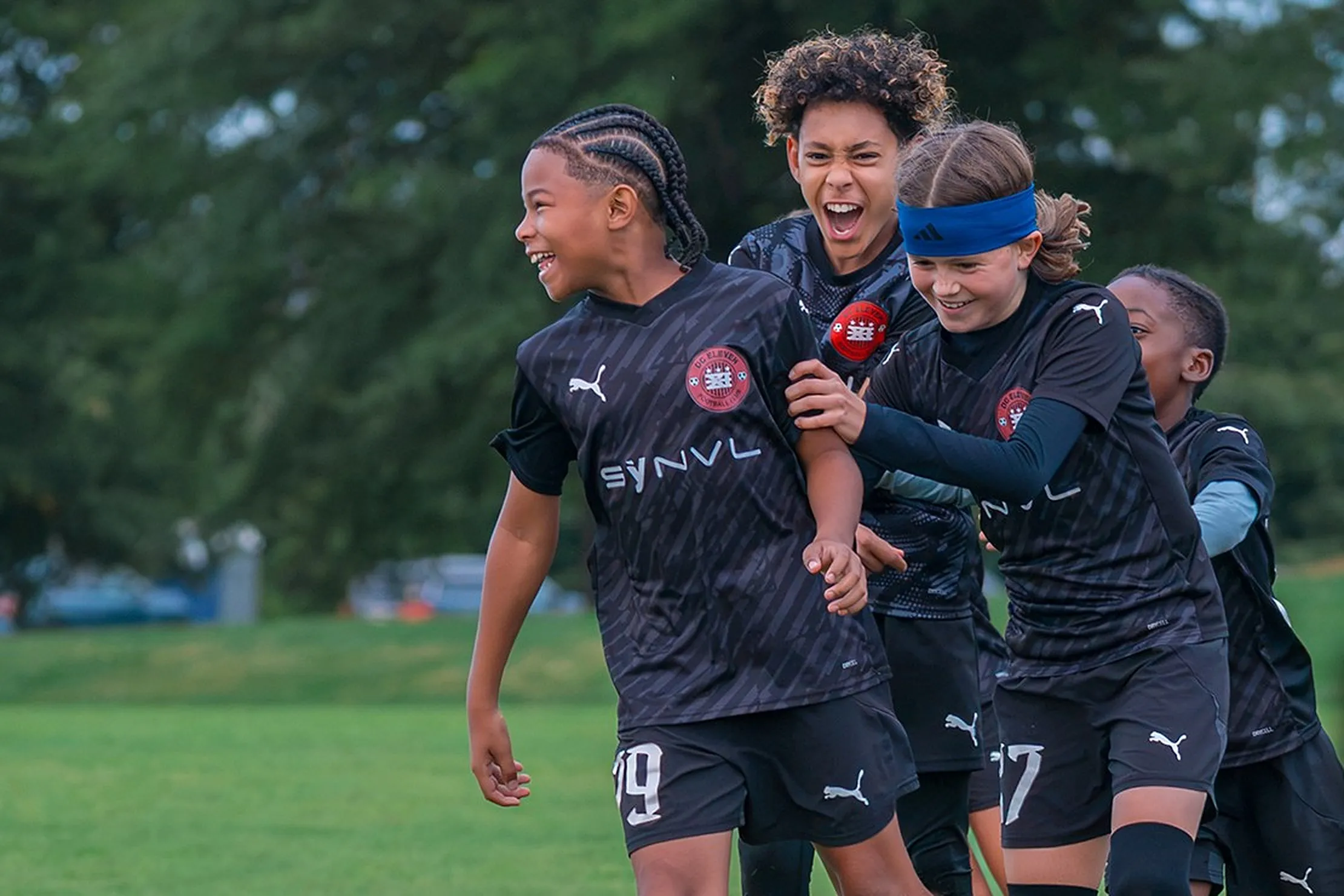 Three smiling players in black DC Eleven kits celebrate together on a grass field.