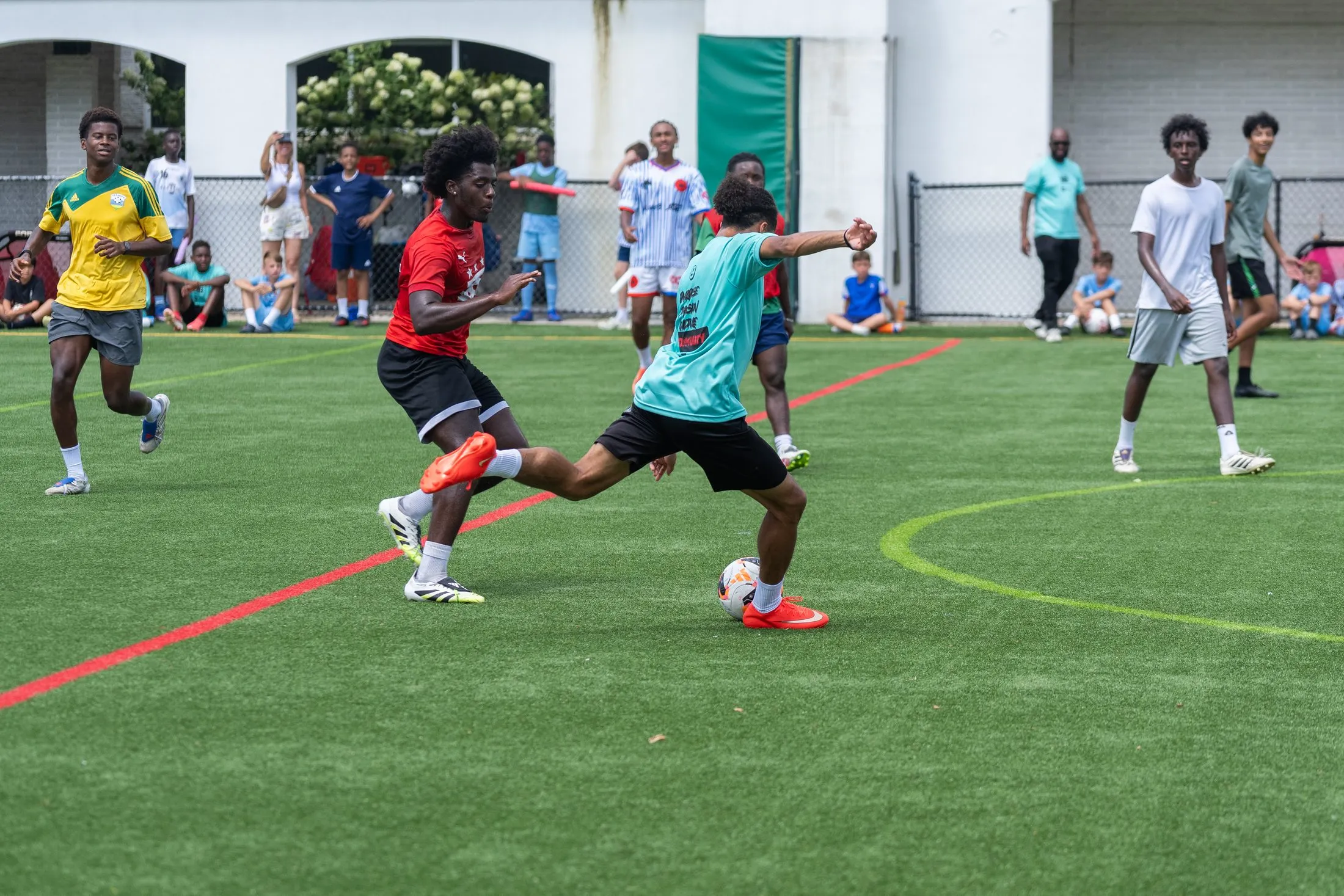A player in a teal shirt strikes the ball while a defender in red closes in, with teammates and onlookers behind them.