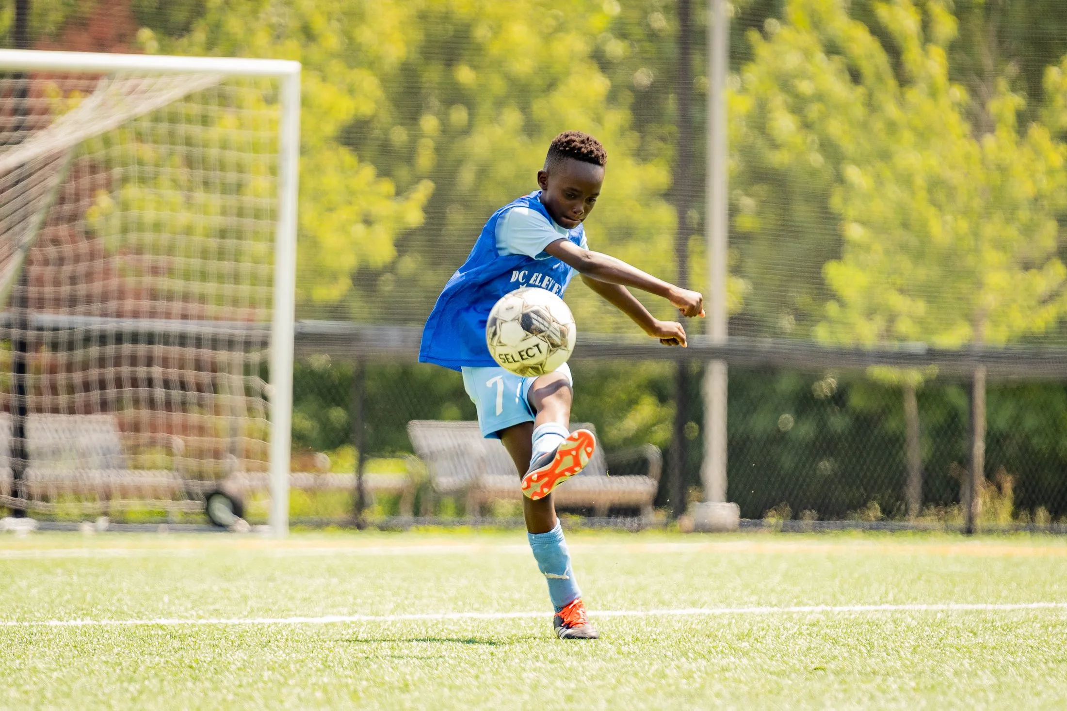 A camper in a blue bib kicks a ball in front of a goal.