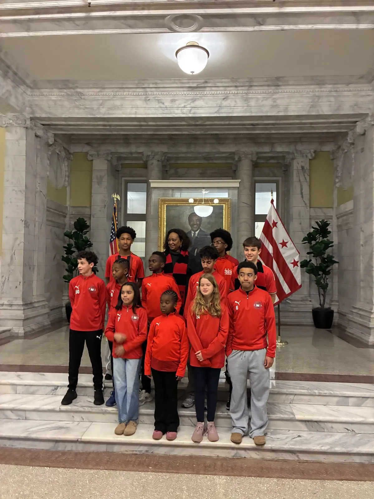 A group of DC Eleven players in red club jackets pose with a woman in a red and black scarf in a marble hall, with the DC flag behind them.