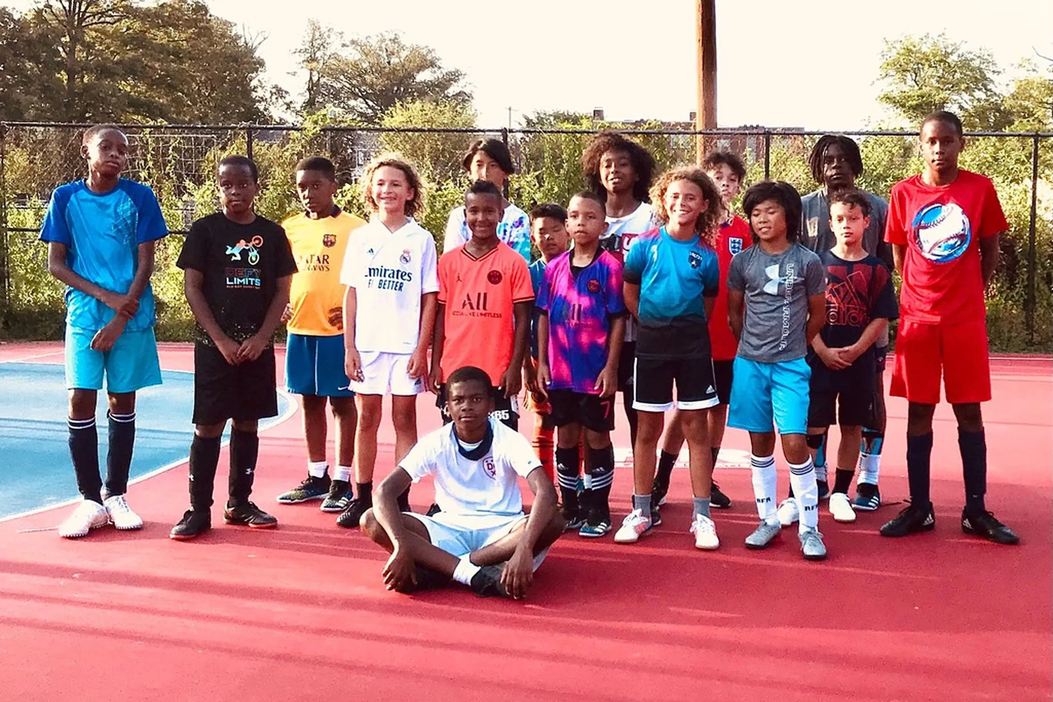 A group of young players in colorful jerseys pose together on a red neighborhood court.