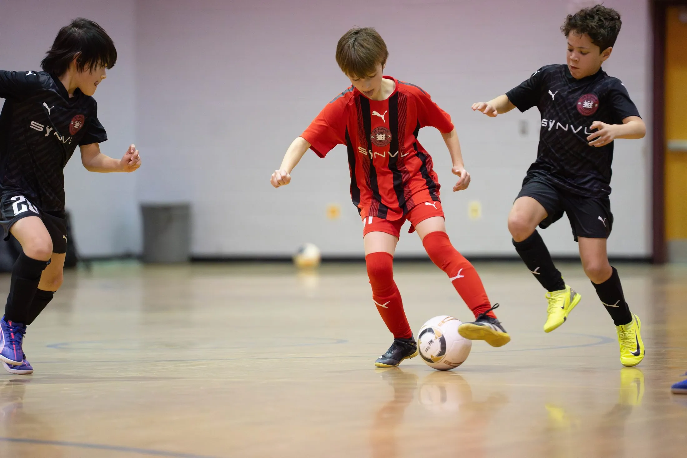 Two players in red and black kits compete for the ball on an indoor gym floor.