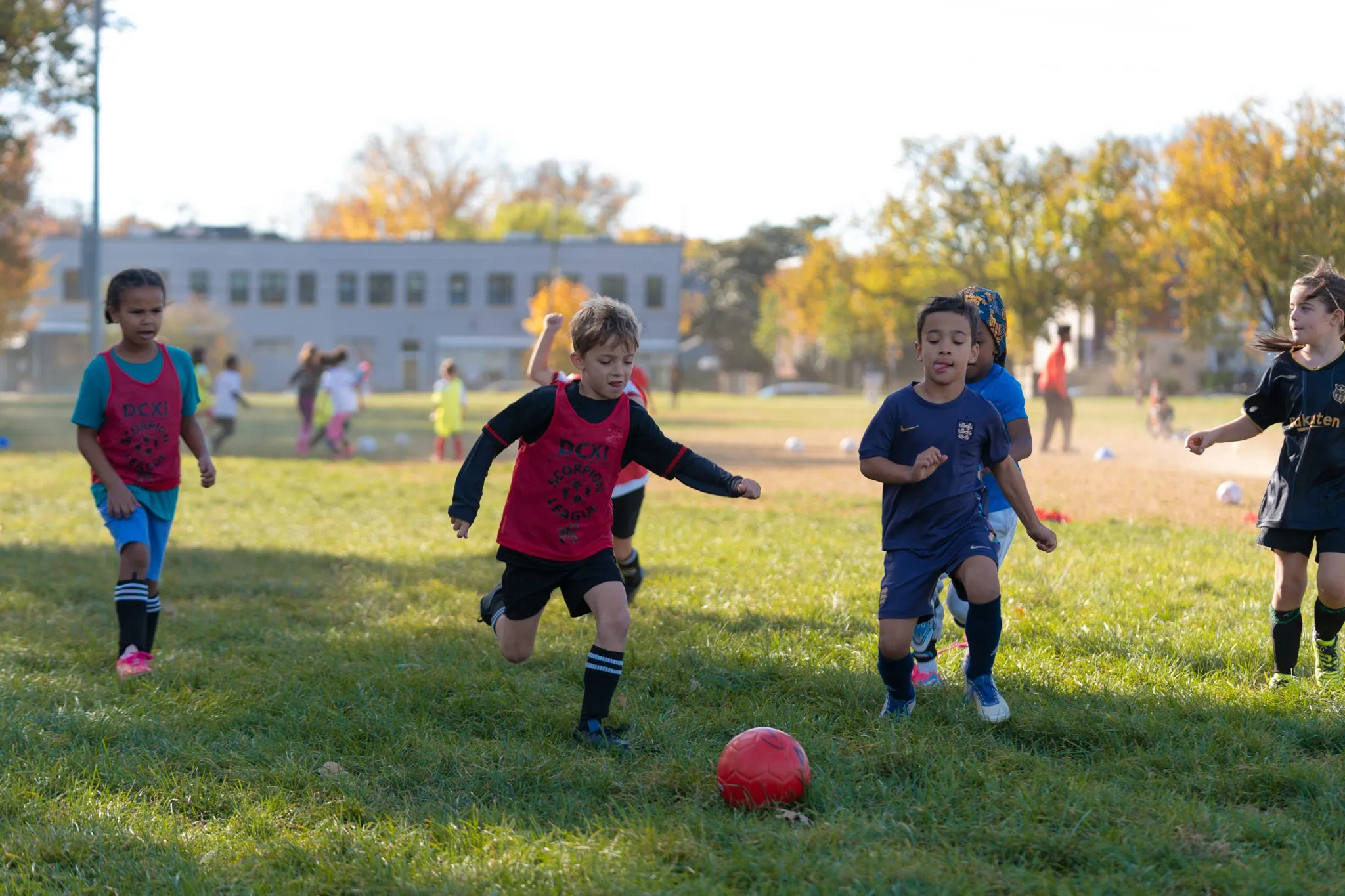 Young players in red and blue jerseys run toward a red ball on a grass field.