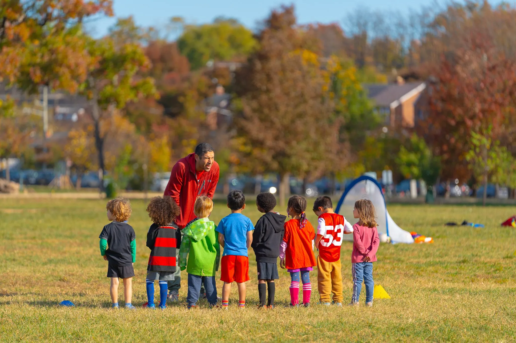 Little players stand in a line on a grass field while a coach crouches beside them.