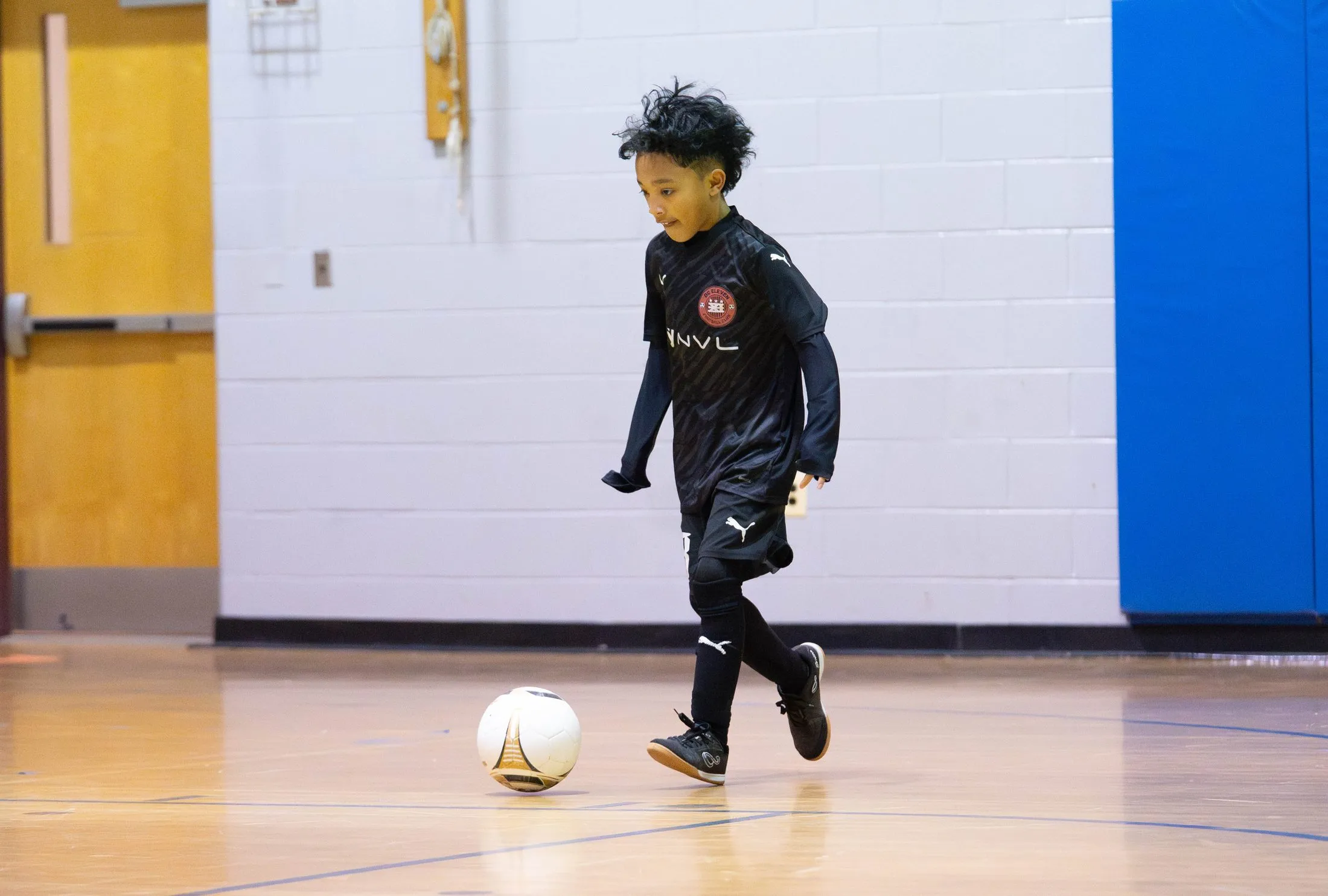 A young player in a black kit dribbles a ball across a gym floor.