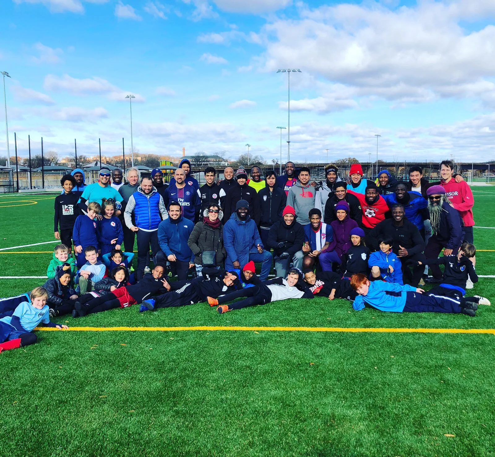 A big group photo of players of many ages in blue jackets, lying and standing together on a turf field.