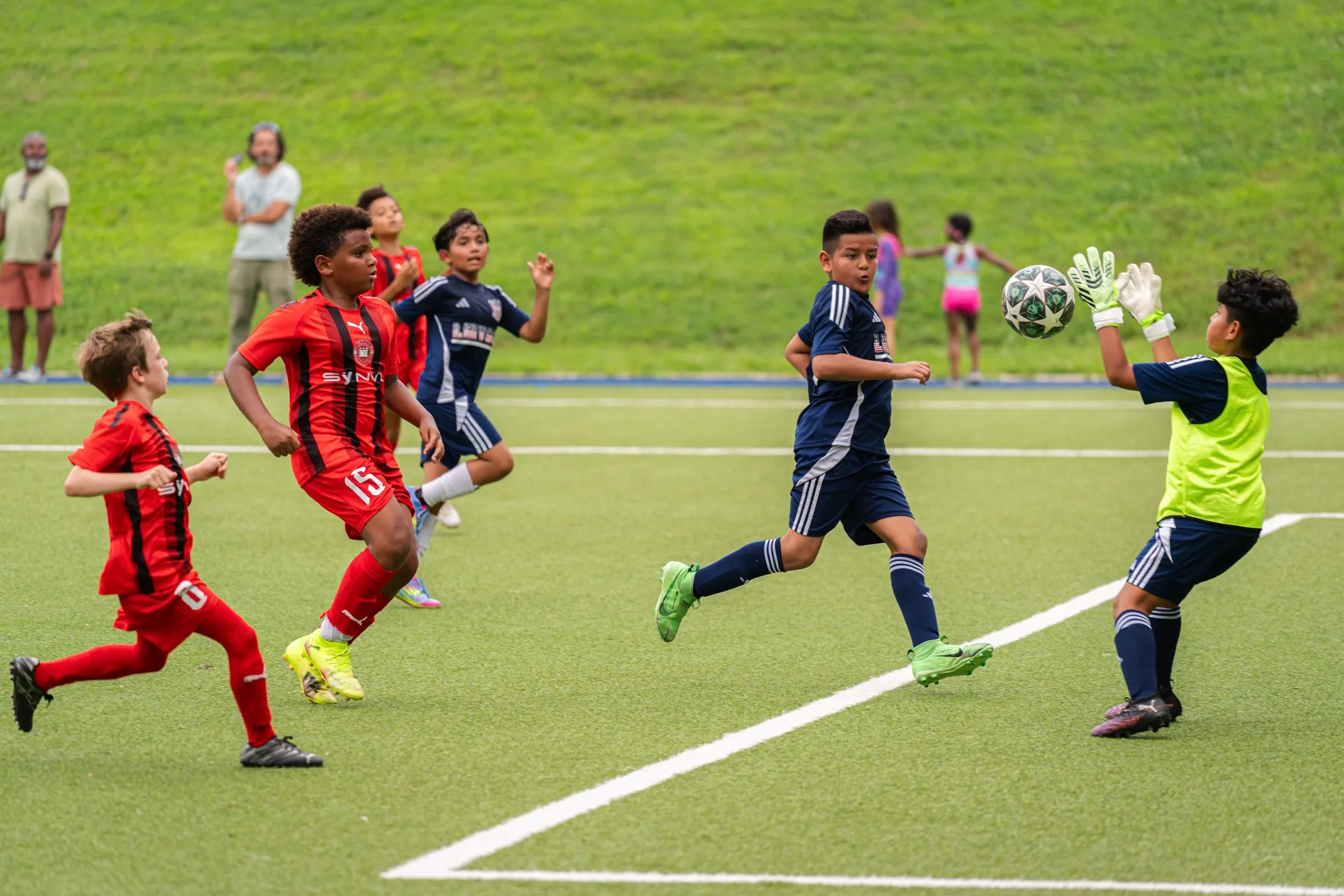 Players in red and navy kits chase a loose ball while a goalkeeper in a green bib reaches for it.